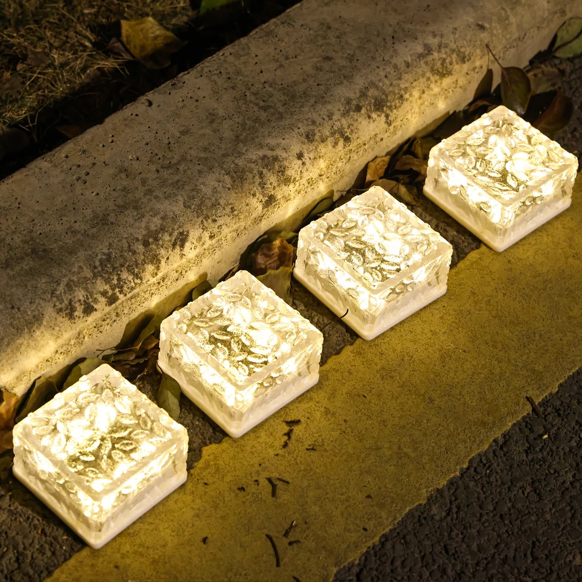 Four solar-powered yard brick lights with a textured glass-like finish are lined up along a sidewalk, emitting a warm white glow.