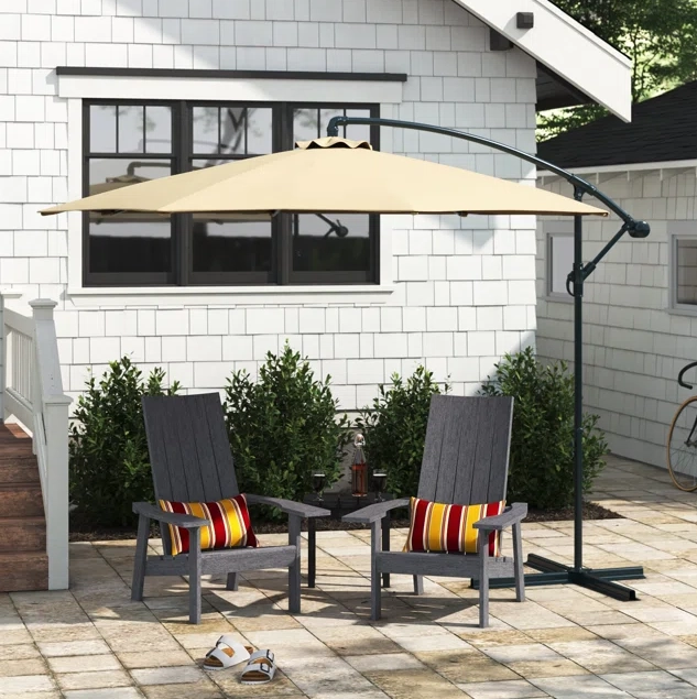 A beige cantilever umbrella with a curved black pole over two grey patio chairs with red-striped cushions and a matching small table.