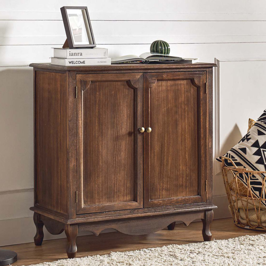 A wooden cabinet with two doors sits against a wall, topped with books and a small decor item. A floor lamp stands beside it, and a basket with patterned pillows is on the floor.