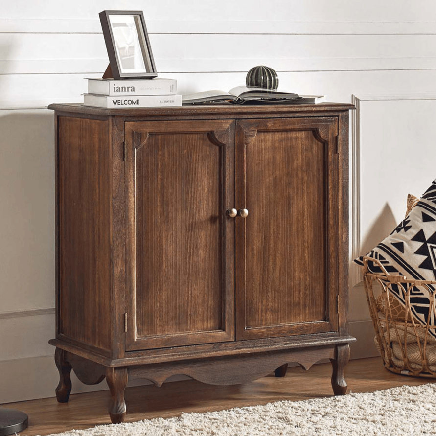 A wooden cabinet with two doors sits against a wall, topped with books and a small decor item. A floor lamp stands beside it, and a basket with patterned pillows is on the floor.