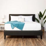 A 12\“ medium plush full mattress is set on a dark bed frame, topped with white bedding and pillows. A teal blanket is draped partially over the bed. A small white nightstand with a lamp sits on one side, and a large potted plant stands beside it.