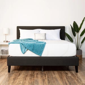 A 12\“ medium plush full mattress is set on a dark bed frame, topped with white bedding and pillows. A teal blanket is draped partially over the bed. A small white nightstand with a lamp sits on one side, and a large potted plant stands beside it.