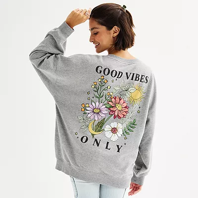 A woman wears a gray oversized sweatshirt with a colorful \“GOOD VIBES ONLY\“ floral design on the back.