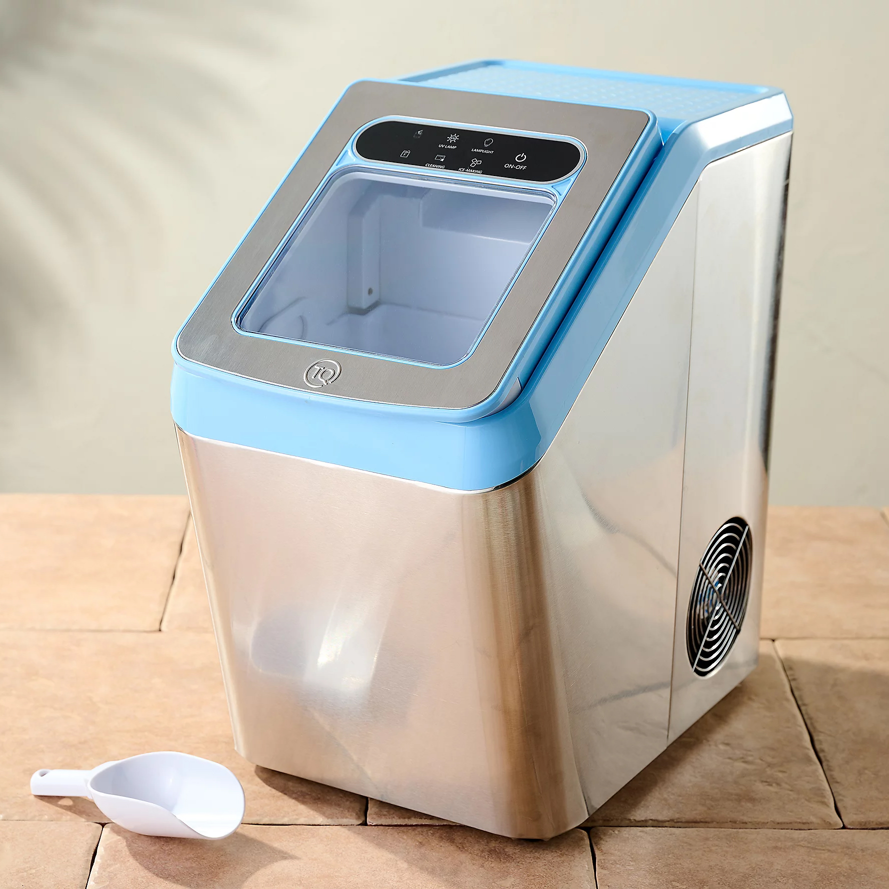 A compact, modern ice maker with a stainless steel body and a blue top accent. Features a transparent lid, control panel, side ventilation fan, and a white ice scoop.
