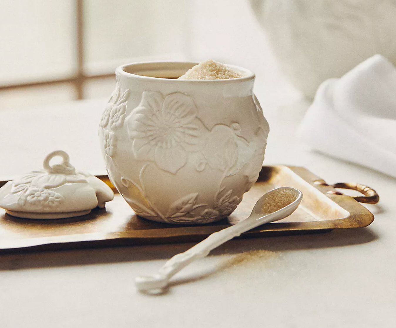 A white ceramic sugar bowl with embossed floral patterns is on display, accompanied by a matching lid and a small spoon on a brass tray.