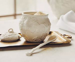 A white ceramic sugar bowl with embossed floral patterns is on display, accompanied by a matching lid and a small spoon on a brass tray.