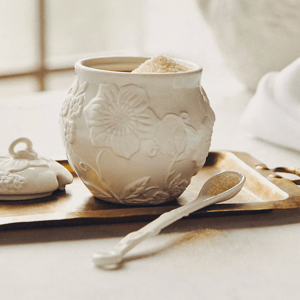 A white ceramic sugar bowl with embossed floral patterns is on display, accompanied by a matching lid and a small spoon on a brass tray.