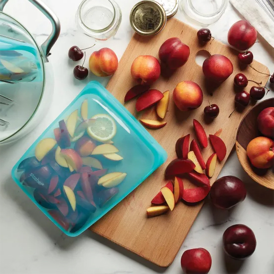 A blue reusable food storage bag lies open with sliced apples and a lemon slice inside, amidst a scattered arrangement of whole and cut peaches and cherries on a kitchen surface.
