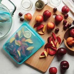 A blue reusable food storage bag lies open with sliced apples and a lemon slice inside, amidst a scattered arrangement of whole and cut peaches and cherries on a kitchen surface.
