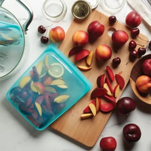 A blue reusable food storage bag lies open with sliced apples and a lemon slice inside, amidst a scattered arrangement of whole and cut peaches and cherries on a kitchen surface.