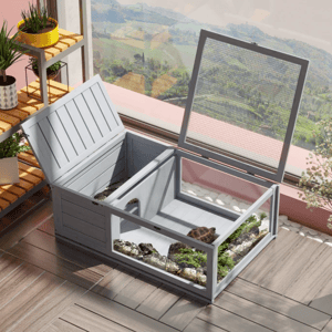 An indoor tortoise enclosure with a mesh lid and clear paneling is placed on a tiled floor, surrounded by potted plants near a window offering a scenic view.