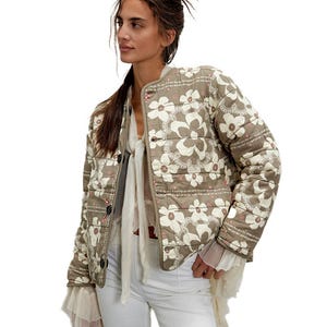 A woman models a quilted floral-patterned jacket paired with a sheer blouse and white pants.