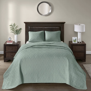 A bedroom setup featuring a bed with a dark wood headboard and a sage green quilted bedspread with matching pillows. Two nightstands, one with a lamp and the other with books and a clock, flank the bed. A round mirror hangs above the bed.