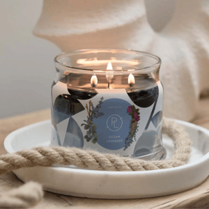 A glass jar candle with three wicks bears the label \“Ocean Lavender\“ and features floral designs. It is placed on a white tray with a decorative rope and emits a warm glow.