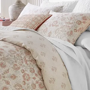 A bed with a patterned comforter and pillowcases featuring floral designs in beige and soft pink tones, complemented by a decorative geometric cushion.