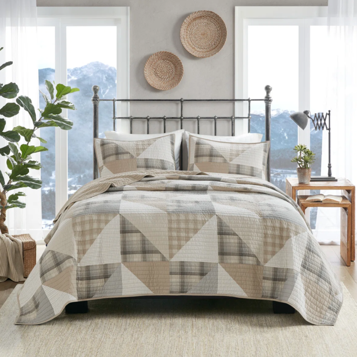 A bed with a geometric quilt and matching pillow shams is shown, featuring a neutral color palette. The room includes woven wall decor, a metal bed frame, wooden side table with books and a lamp, a potted plant, and large windows with a mountain view.