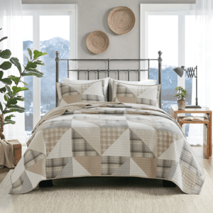 A bed with a geometric quilt and matching pillow shams is shown, featuring a neutral color palette. The room includes woven wall decor, a metal bed frame, wooden side table with books and a lamp, a potted plant, and large windows with a mountain view.