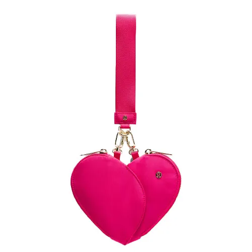 A bright pink, heart-shaped wristlet with dual zippered compartments, a gold clasp, and a detachable strap featuring the Lululemon logo.