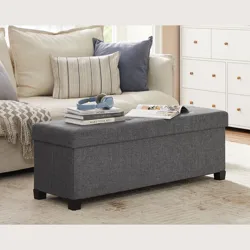 A grey upholstered storage bench with black legs is placed in a living room next to a beige sofa with cushions. It holds books, headphones, and a magazine, with a dresser visible in the background.