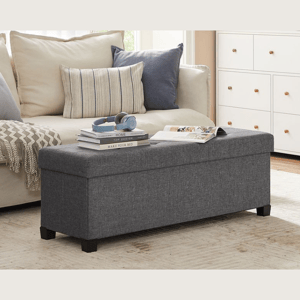 A grey upholstered storage bench with black legs is placed in a living room next to a beige sofa with cushions. It holds books, headphones, and a magazine, with a dresser visible in the background.
