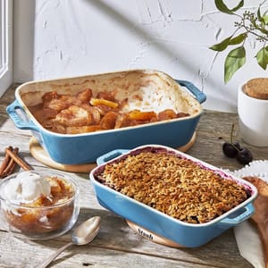 The 2-piece Staub bakeware set, in blue, includes a deep baking dish filled with spiced apples and a shallower dish topped with a golden oat crumble, accompanied by a bowl of the apple dessert with ice cream.
