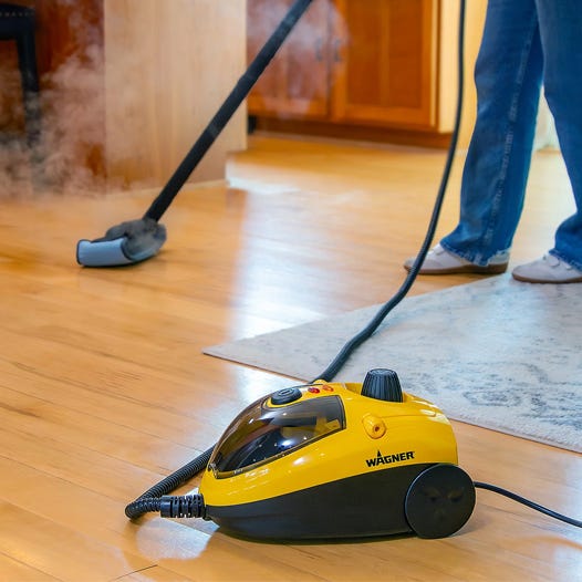 A Wagner steam cleaner is in use on a hardwood floor, releasing steam for cleaning purposes. A person is holding the hose, wearing blue jeans and sneakers.