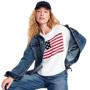 A woman wears a white sweater with an American flag design, paired with a denim jacket and jeans. She also has a dark cap.