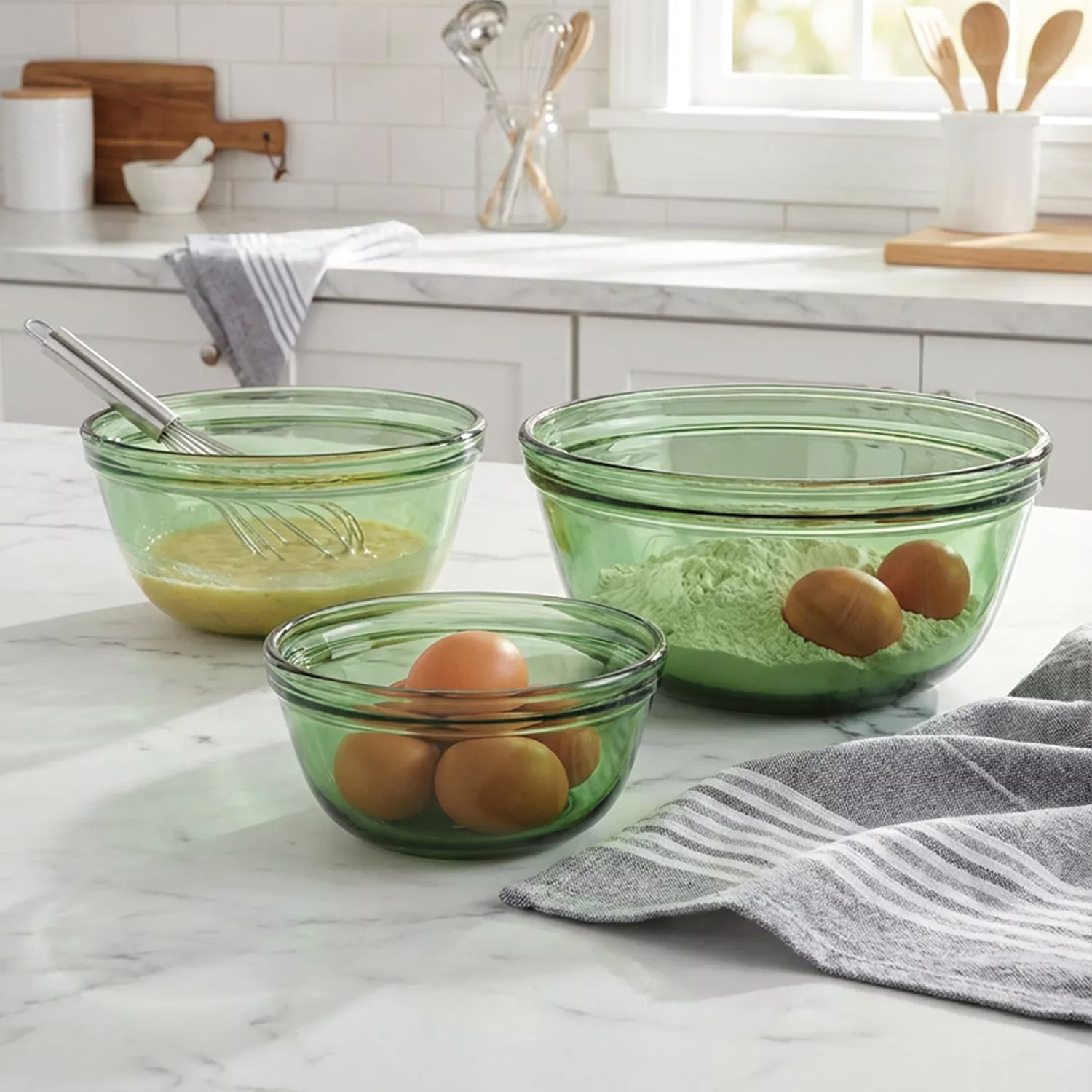 Three green glass mixing bowls of varying sizes contain eggs, flour, and a whisk with batter. A gray and white striped towel is nearby on a marble countertop in a kitchen setting.