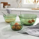 Three green glass mixing bowls of varying sizes contain eggs, flour, and a whisk with batter. A gray and white striped towel is nearby on a marble countertop in a kitchen setting.