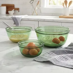 Three green glass mixing bowls of varying sizes contain eggs, flour, and a whisk with batter. A gray and white striped towel is nearby on a marble countertop in a kitchen setting.