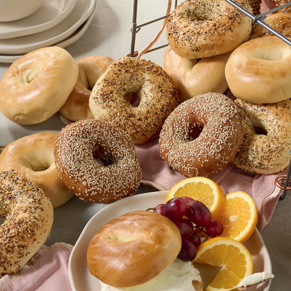 A variety of bagels, including plain, sesame, and everything bagels, are arranged on a table with plates, cream cheese, orange slices, and grapes.