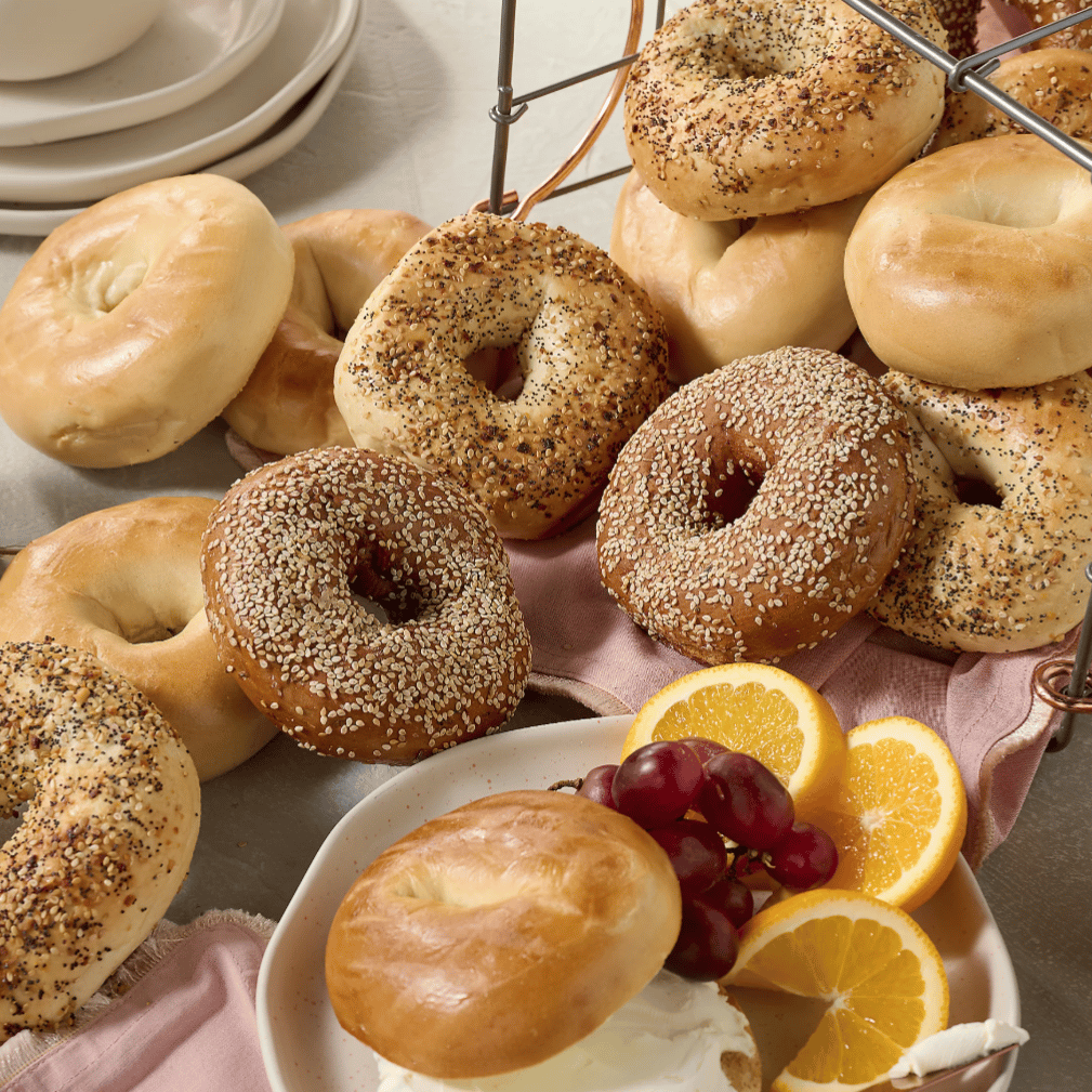 A variety of bagels, including plain, sesame, and everything bagels, are arranged on a table with plates, cream cheese, orange slices, and grapes.