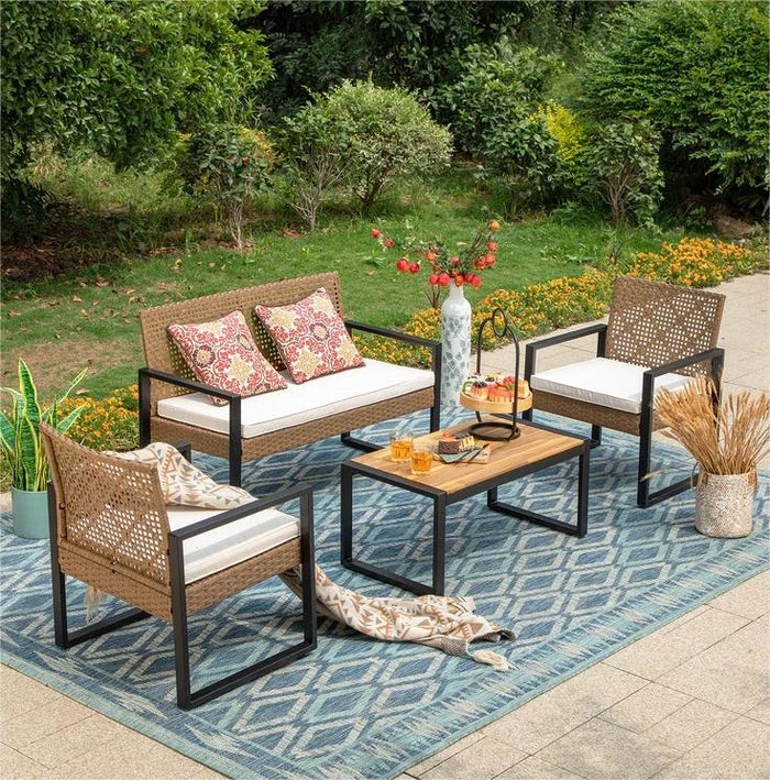 A stylish outdoor furniture set featuring a loveseat, two armchairs, and a coffee table on a blue patterned rug. The seating has wicker design with white cushions, and the table holds a vase and snacks. The backdrop includes greenery and flowers.