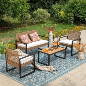 A stylish outdoor furniture set featuring a loveseat, two armchairs, and a coffee table on a blue patterned rug. The seating has wicker design with white cushions, and the table holds a vase and snacks. The backdrop includes greenery and flowers.