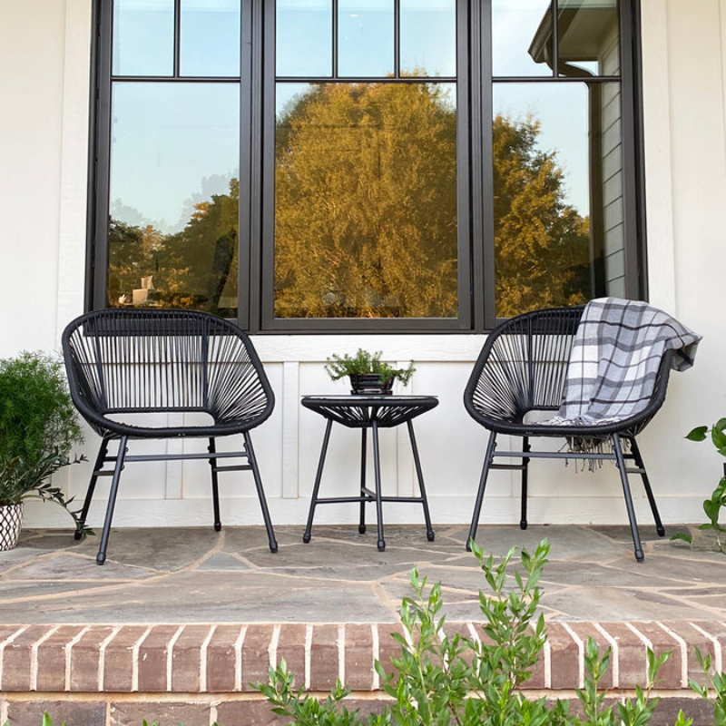 The 3-piece Phi Villa Outdoor Wicker Set includes two black, rounded wicker chairs with armrests and a small round side table. One chair features a gray plaid blanket for a cozy touch, and the set is placed on a stone patio with brick steps.