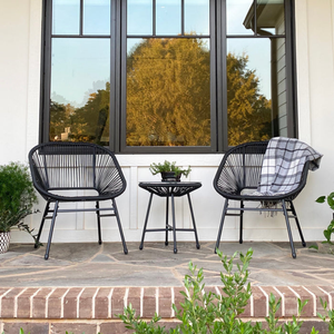 The 3-piece Phi Villa Outdoor Wicker Set includes two black, rounded wicker chairs with armrests and a small round side table. One chair features a gray plaid blanket for a cozy touch, and the set is placed on a stone patio with brick steps.