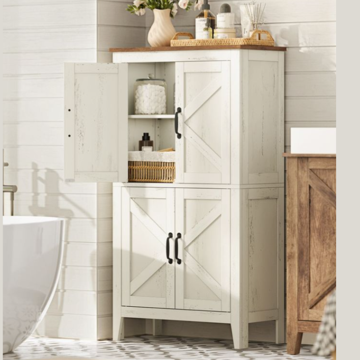 A white wooden cabinet with black handles, featuring an X-design, stands in a bathroom setting. It has three compartments with shelves, one door open revealing toiletries, and decor items like flowers and soap dispensers on top.
