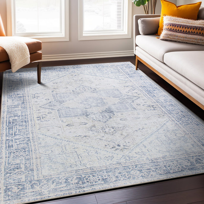 An 8' x 10' machine-washable area rug with a light blue and white intricate pattern, placed in a bright room with a sofa and chair nearby.