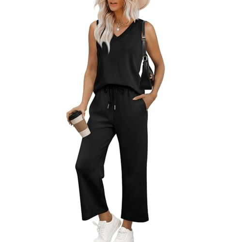 A woman is wearing a sleeveless black jumpsuit with a V-neck and drawstring waist, paired with white sneakers and a black shoulder bag. She's holding a coffee cup and wearing a wide-brimmed hat.