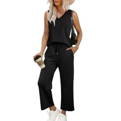 A woman is wearing a sleeveless black jumpsuit with a V-neck and drawstring waist, paired with white sneakers and a black shoulder bag. She's holding a coffee cup and wearing a wide-brimmed hat.