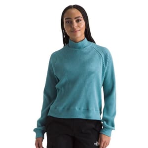 A person is wearing a turquoise waffle-knit turtleneck sweater paired with black pants featuring \“The North Face\“ logo.