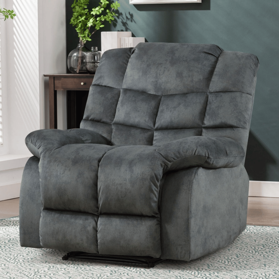 A plush, gray reclining armchair is set against a dark green wall, with a light rug beneath it and a small wooden side table with books and decorative items nearby.