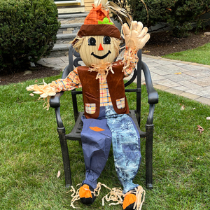 A decorative scarecrow figure is seated on a metal chair, wearing a plaid shirt, a brown vest, and blue jeans, with straw accents.