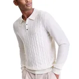 A man wears a cream-colored, cable-knit sweater with a collar and button placket, paired with light-colored trousers.