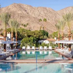 A resort pool area is surrounded by white lounge chairs and umbrellas, with tall palm trees and a mountainous backdrop.