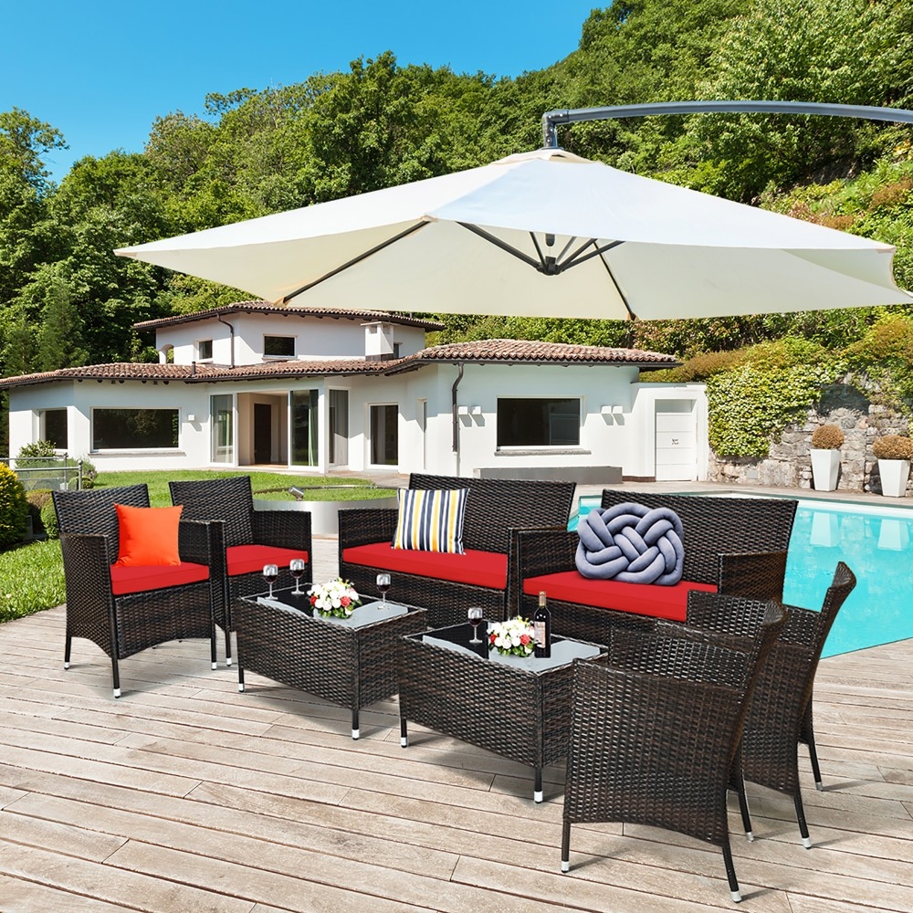 The 8pc Patio Conversation Set includes wicker seating with red cushions: a loveseat, two armchairs, and four stools. It features two glass-top coffee tables and is covered by a large white umbrella, set on a wooden deck by a pool.