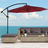 A patio setup with a red cantilever umbrella shading a wicker sofa set against a sea backdrop, featuring cushions and a glass table with a teapot, books, and a plant.