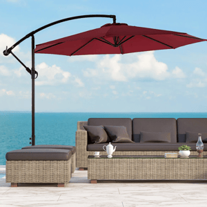 A patio setup with a red cantilever umbrella shading a wicker sofa set against a sea backdrop, featuring cushions and a glass table with a teapot, books, and a plant.