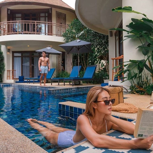 A woman relaxes by a pool while reading a book, wearing sunglasses and a bikini, with a man standing in the background. The setting features an upscale villa with blue poolside loungers and umbrellas.