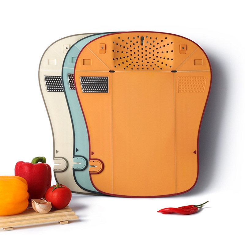 Three stackable cutting boards with integrated features like grater, slicer, and juice drainer, shown in orange, blue, and beige. They are displayed alongside vegetables like bell peppers, a tomato, and garlic on a small wooden board.